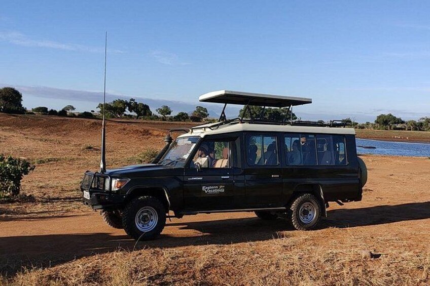 A black safari vehicle with a roof rack and a spare tire mounted on the back, parked on a dirt road with dry grass and a body of water in the background. 4