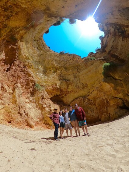 A group of people standing in a sandy area with a large rock formation and a blue sky above. 5