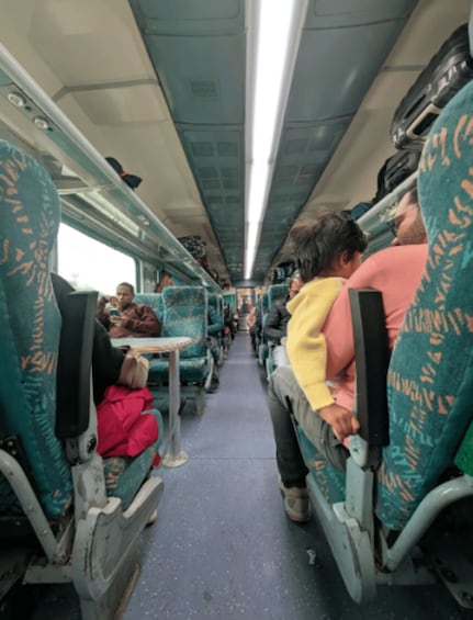 A train interior with passengers seated, green patterned seats, overhead compartments, and a central aisle. 3