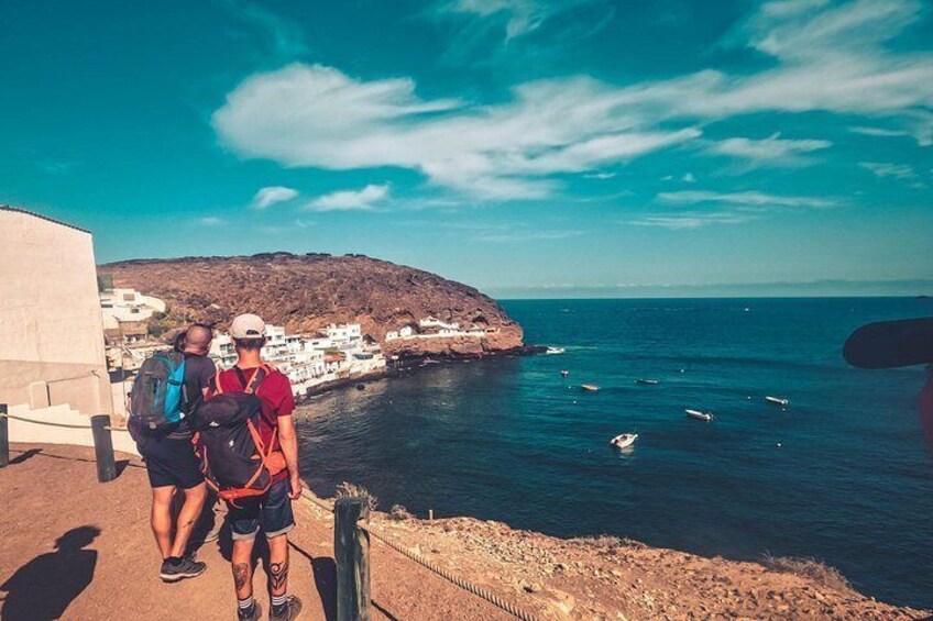 Two people standing on a cliff overlooking a coastal town with white buildings and a clear blue sky. 3