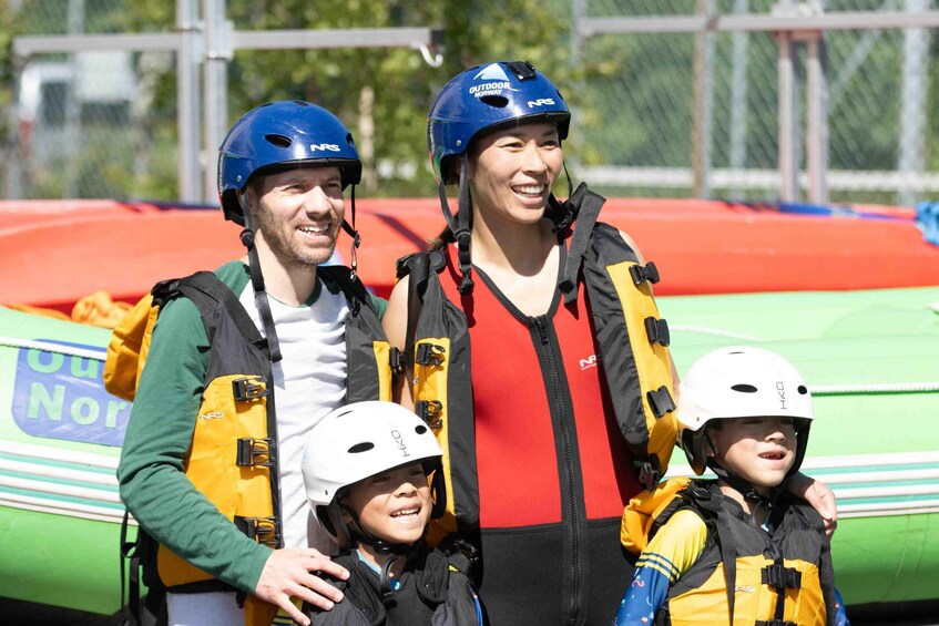 Three people wearing helmets and life jackets, standing in front of a green inflatable raft. 1