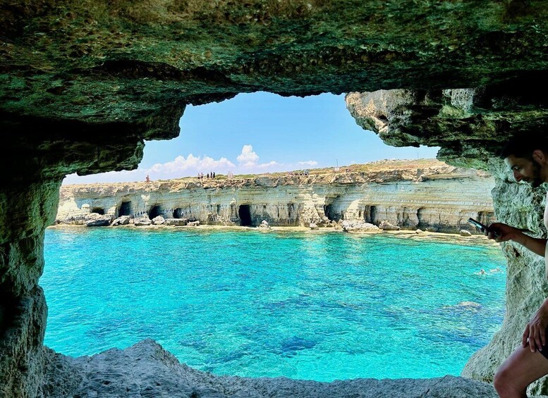 Une personne se trouve à l’intérieur d’une grotte et observe un paysage côtier avec une eau bleue et limpide et des falaises rocheuses. 1