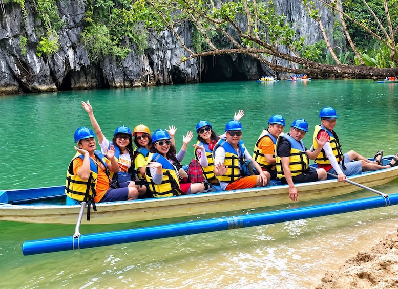 A group of people in a boat wearing life jackets and helmets, paddling through a calm river with rocky cliffs and greenery in the background. 2