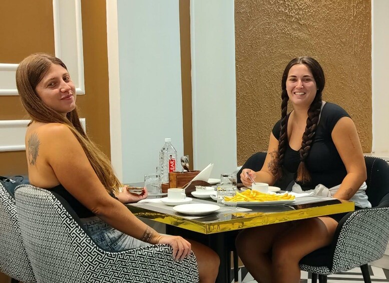 Two women sitting at a table with plates of food, a bottle of water, and a cup. 2