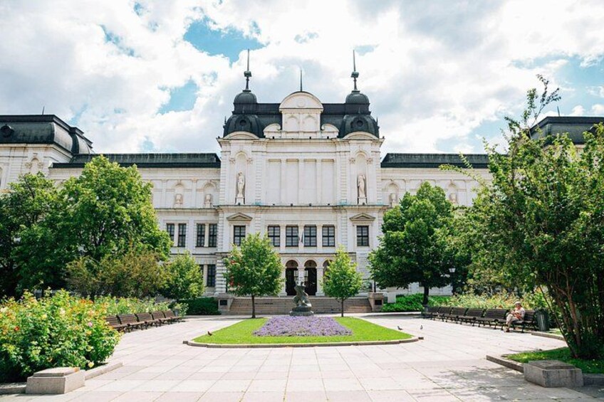 Ein großes, weißes Gebäude mit einer zentralen Kuppel und zwei kleineren Kuppeln, umgeben von einem angelegten Garten mit Bänken. 2