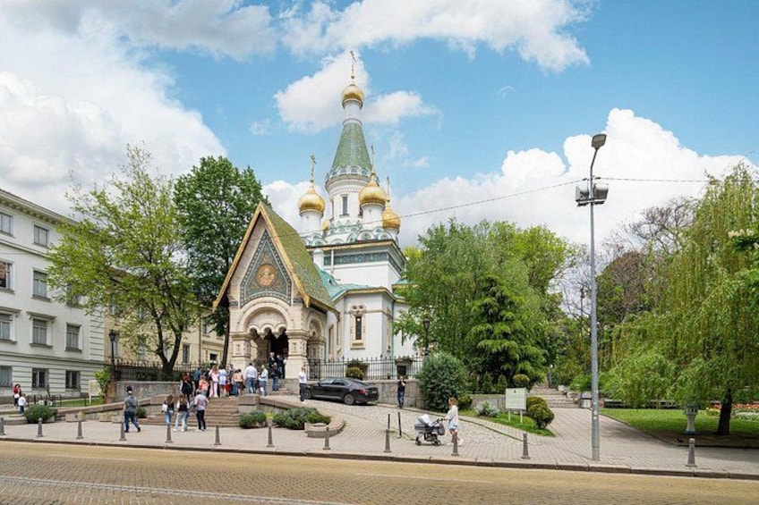 Eine historische Kirche mit goldenen Kuppeln, ein gepflasterter Platz und Menschen, die sich darin bewegen. 1