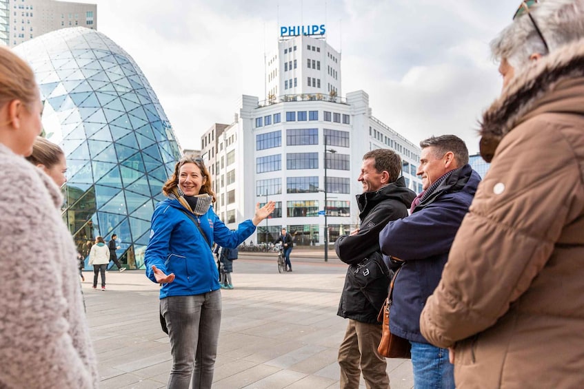 A group of people standing outdoors in an urban setting with modern architecture, including a building with a Philips sign and a distinctive glass dome structure. 3