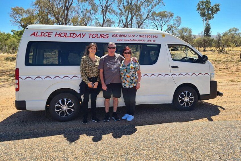 A white van with 'ALICE HOLIDAY TOURS' written on the side, parked on a dirt road with sparse vegetation and trees in the background. 2