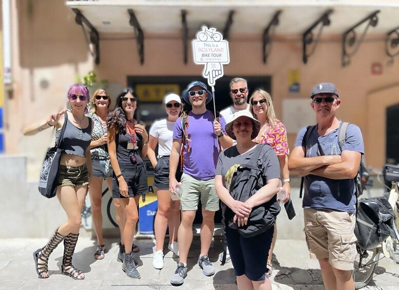 A group of people posing for a photo in front of a building with a sign that reads "This is a Sicilyland Bike Tour." 3