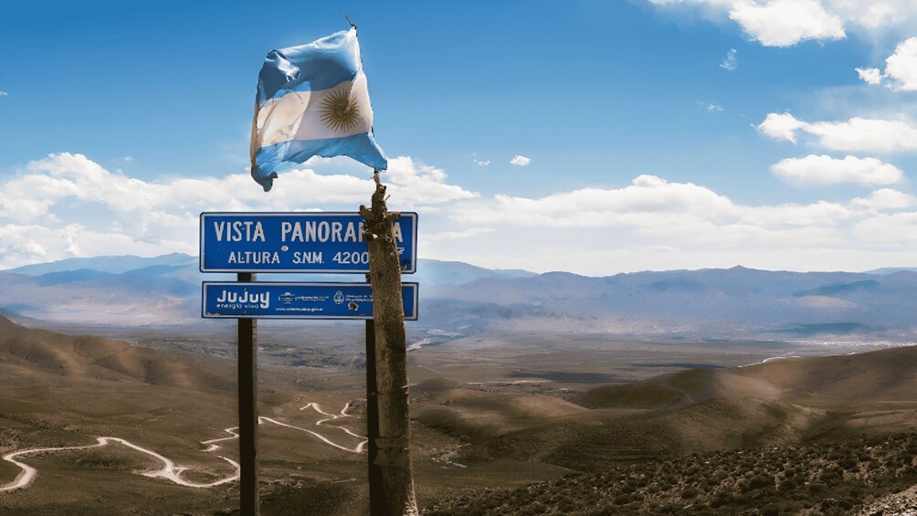A flag, a sign with 'Vista Panorama' and 'Altura S.N.M. 4200', and a vast mountainous landscape. 4