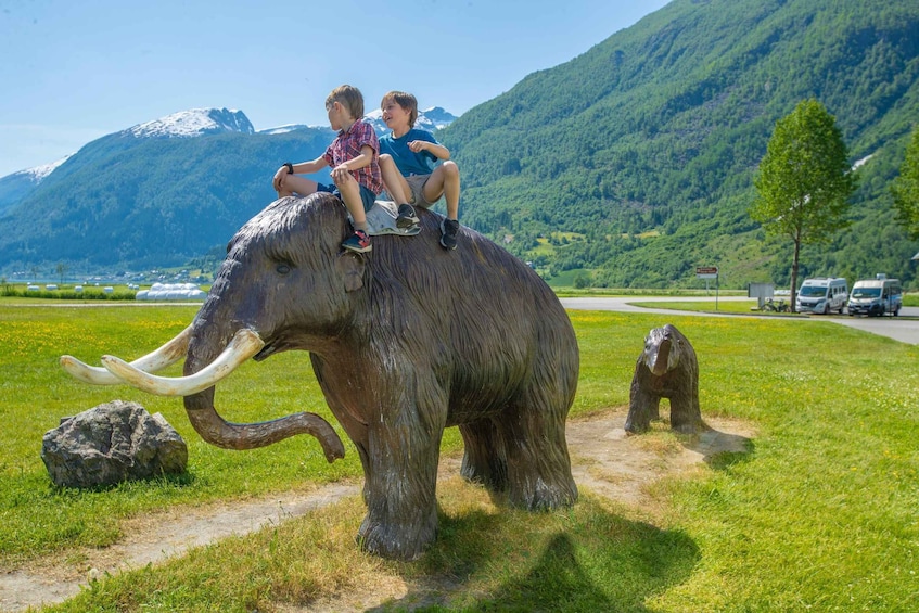 Two people riding a large elephant statue in a grassy field with mountains in the background. 3