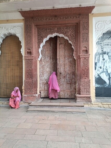 A woman in pink attire seated on steps in front of a intricately carved wooden door with floral and geometric patterns. 2