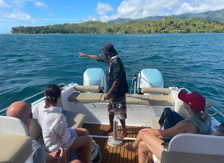 A boat with passengers on a calm sea, a man pointing towards the horizon, and a forested island in the background. 2