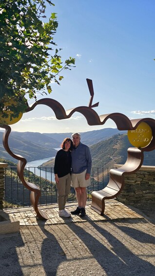 A couple standing in front of a metal sculpture with a scenic view of hills and a body of water in the background. 4