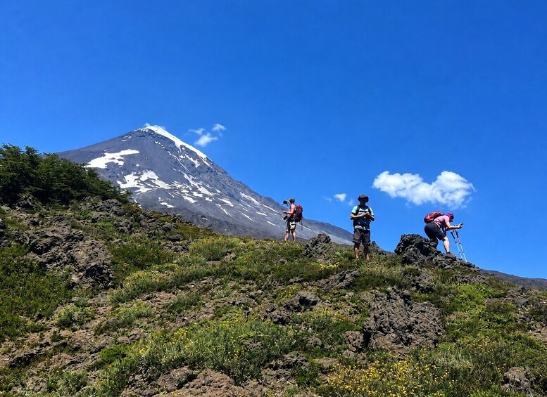 Tres excursionistas en una ladera rocosa de montaña, con un volcán nevado al fondo. 5