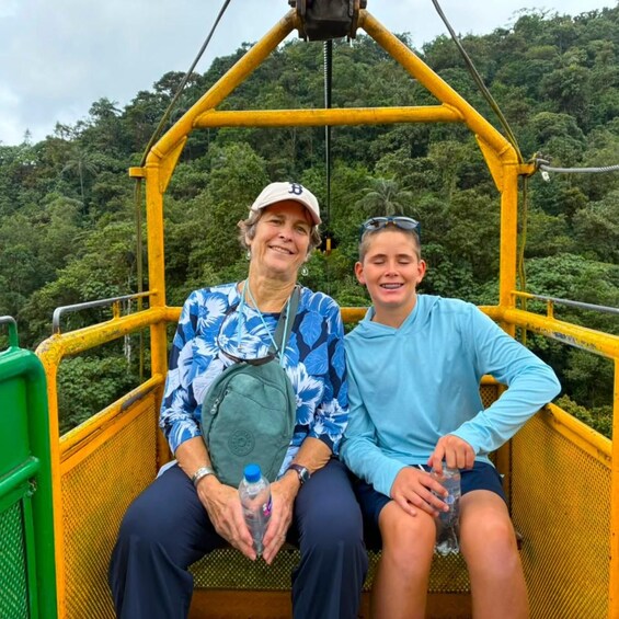 Two people seated on a yellow cable car amidst a lush green forest. 3