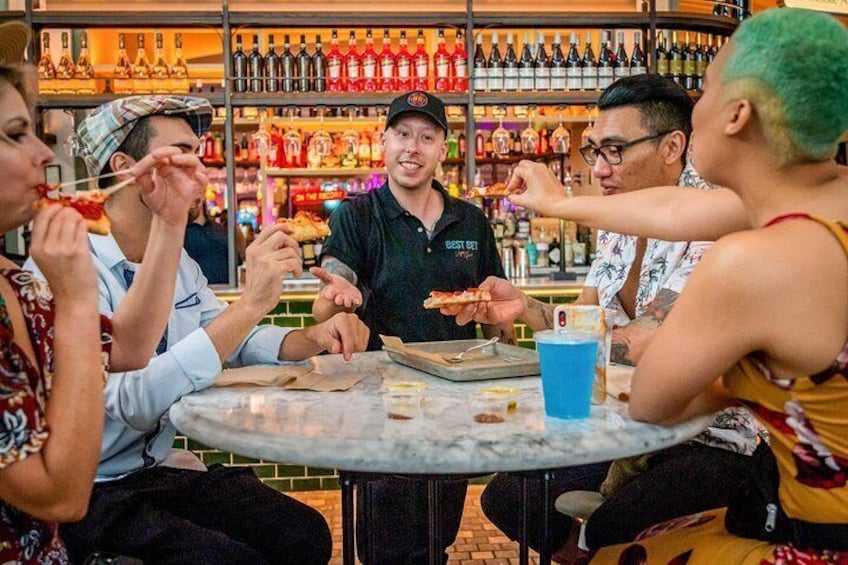 A group of people sitting around a table, eating pizza and drinking from a blue cup. 3