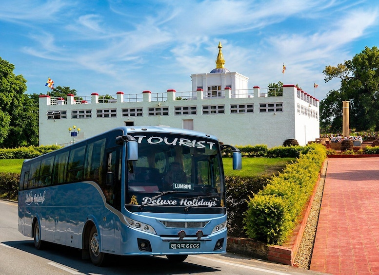 Een blauwe bus met 'Deluxe Holidays' en 'Lumbini' op de voorkant, geparkeerd voor een wit gebouw met een gouden torenspits. 2