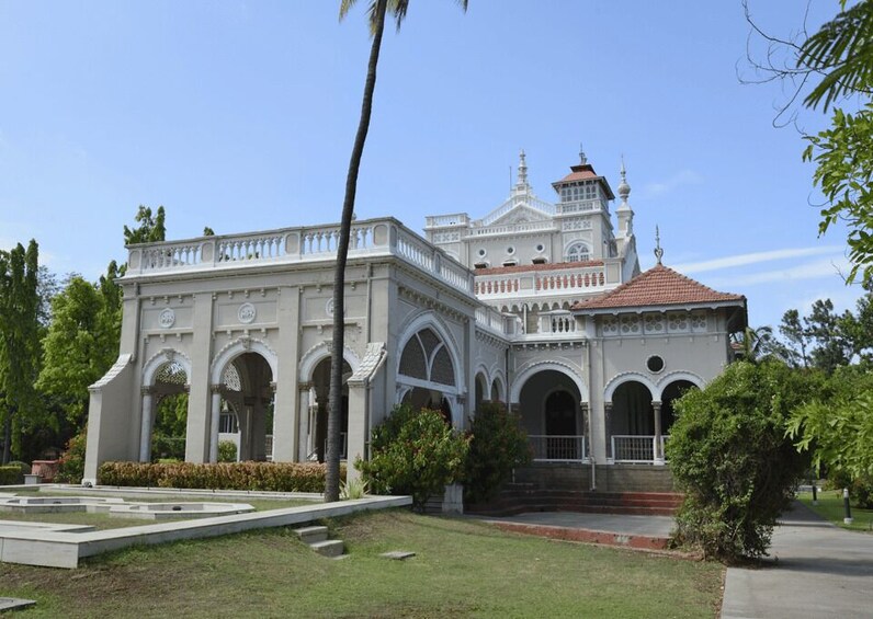 A large, ornate white building with arched windows and a red-tiled roof, surrounded by well-maintained gardens and palm trees. 1