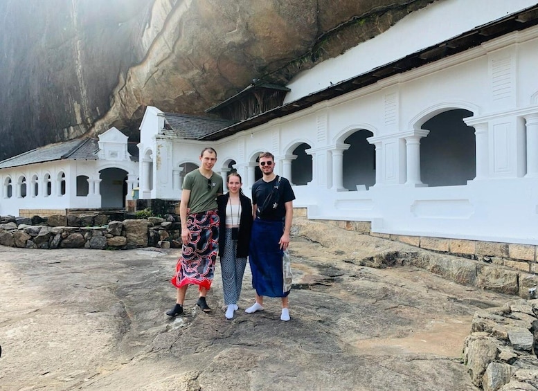 A group of people standing in front of a white building with arched windows and a large rock formation in the background. 1