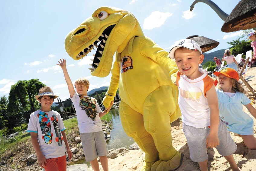 A person in a dinosaur costume with a logo on the chest, standing on a sandy beach with a river and greenery in the background. 1