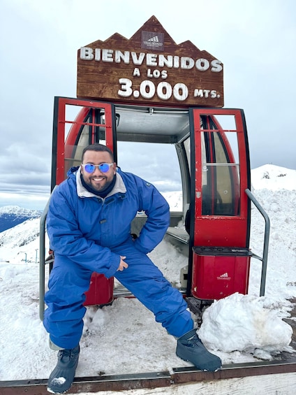 A man in a blue snowsuit posing in front of a red chairlift with a sign that reads "BIENVENIDOS A LOS 3.000 MTS." 1