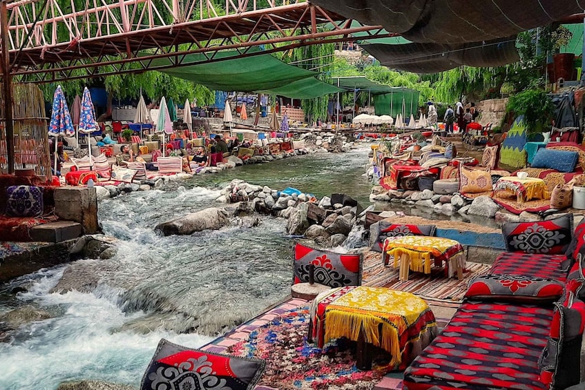 A riverside market with colorful textiles and a bridge overhead. 2