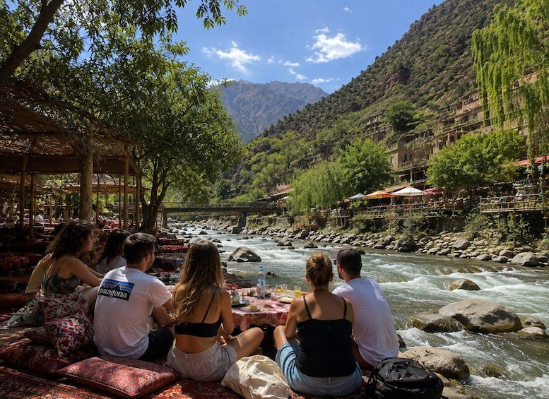 A group of people sitting at a table by a river, with mountains in the background and a bridge crossing the river. 4