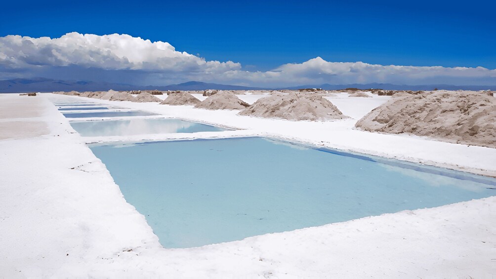 Eine Reihe rechteckiger Pools mit klarem, blauem Wasser, umgeben von weißen Salzflächen und Bergen im Hintergrund. 3
