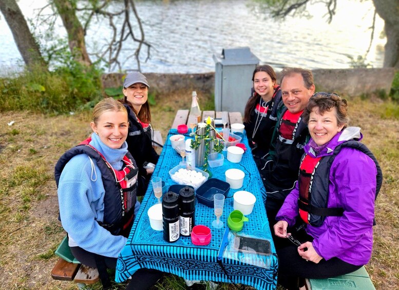A group of people sitting at a picnic table outdoors with a body of water in the background. 2