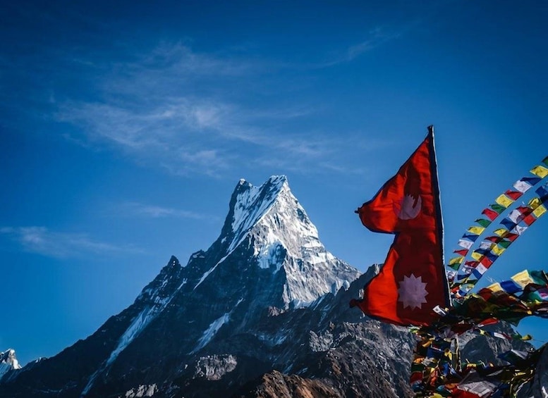 A snow-capped mountain peak under a clear blue sky with a red flag featuring a white emblem and colorful prayer flags in the foreground. 2