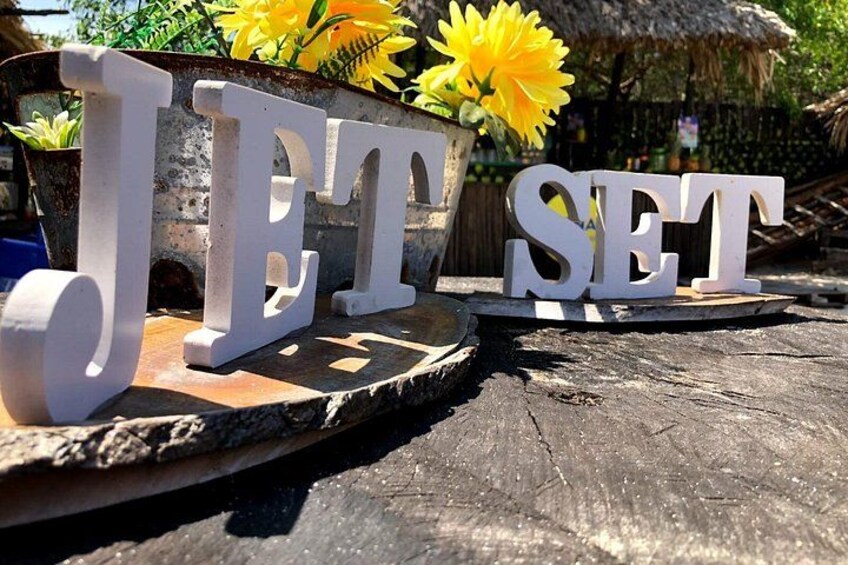 Witte houten letters met de tekst 'JET SET' op een houten oppervlak, met een rieten dak en planten op de achtergrond. 3