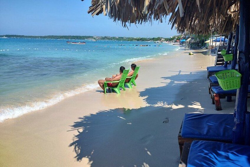 Een strand met twee personen zittend op stoelen, een boot in het water en strandparasols. 5