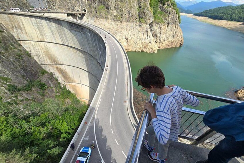 A curved road alongside a dam with a river below, a person standing on a railing, and a blue car on the road. 2