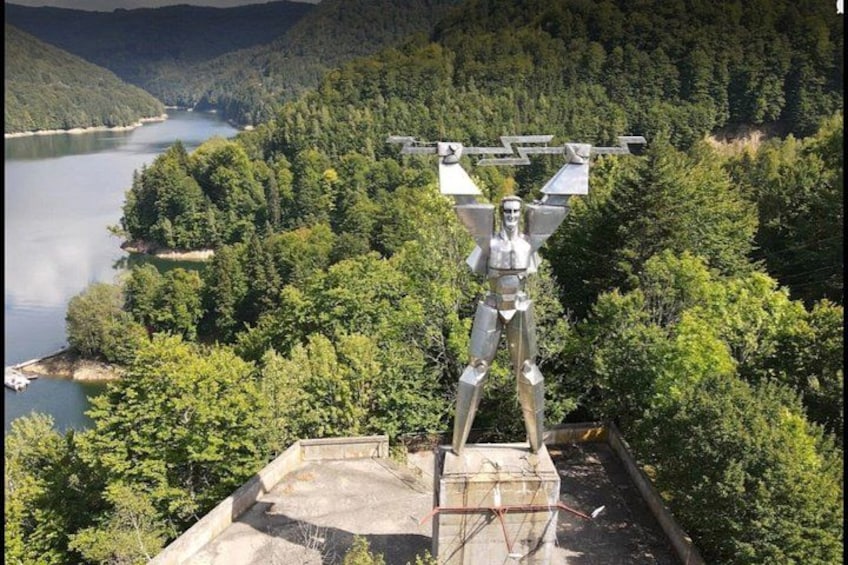 A statue of a humanoid figure on a platform with a lake and forest in the background. 3