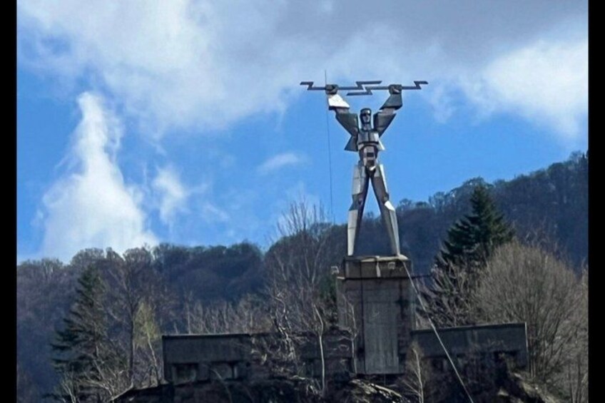 A sculpture of a humanoid figure with a drone on its head, set against a mountainous backdrop. 3