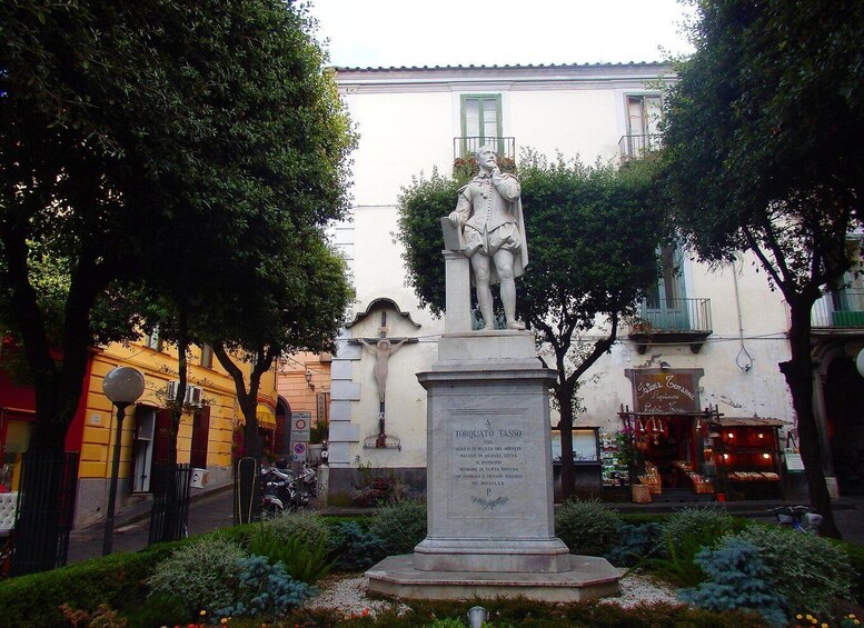 A statue of a man in a white coat with a cross, surrounded by greenery and buildings. 3
