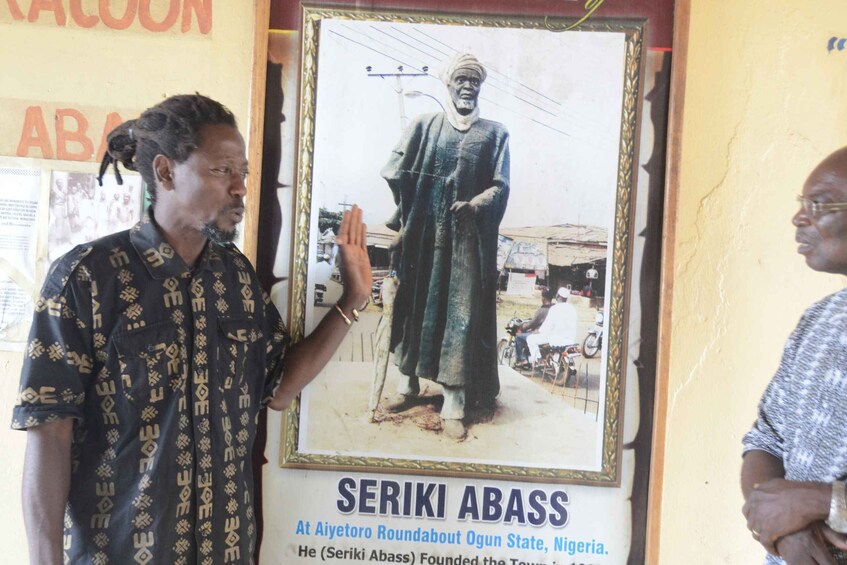 A man standing in front of a poster of Seriki Abass with a caption about him founding the town at Aiyetoro Roundabout, Ogun State, Nigeria. 3
