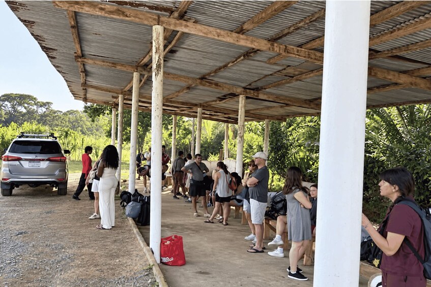 A covered outdoor area with people standing and walking, a parked car, and a red bag on the ground. 2
