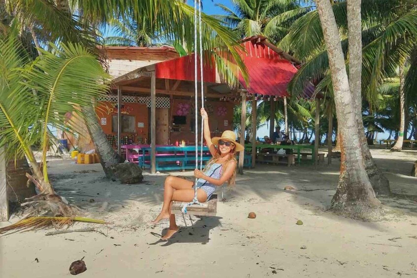 A person sitting on a swing under a palm tree on a sandy beach with a building in the background. 2