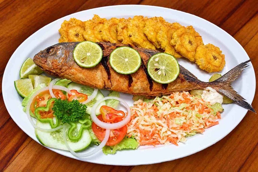 A whole grilled fish with lime slices, a side of coleslaw, and fried plantains on a white plate. 5