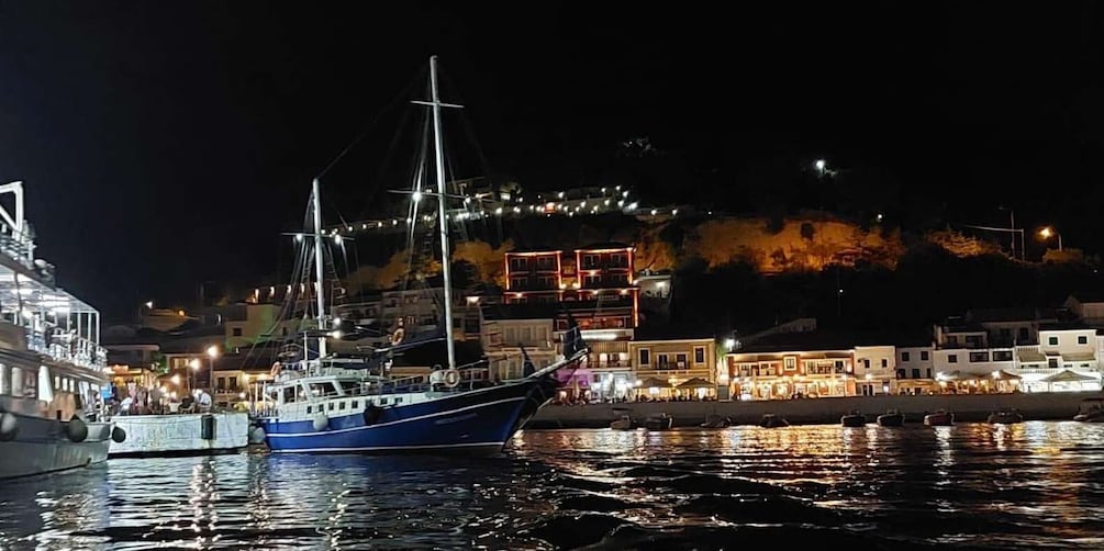 A nighttime harbor with a large sailing ship docked, buildings along the shore, and a hill with lights in the background. 2