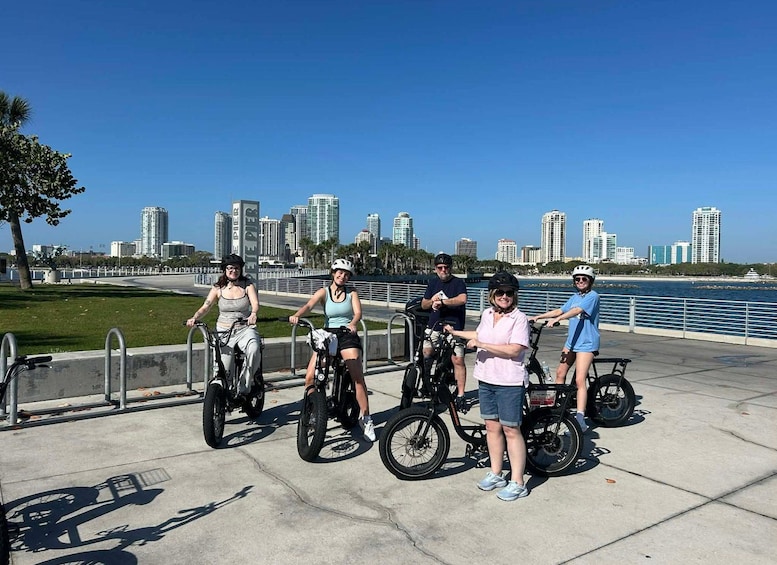 A group of people on bicycles and tricycles by a waterfront with a city skyline in the background. 4