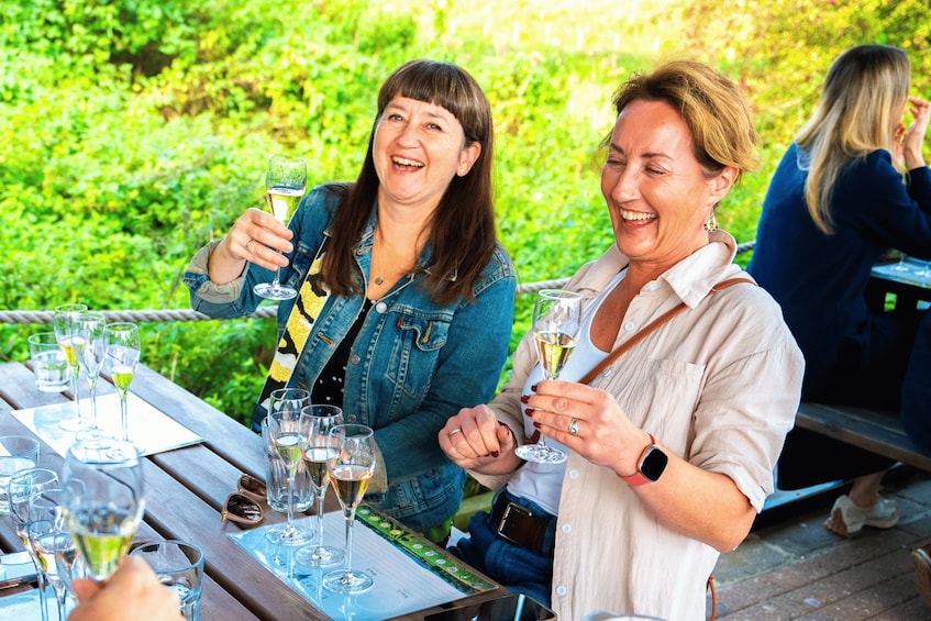 Two women toasting with champagne glasses at an outdoor table with a lush green background. 1