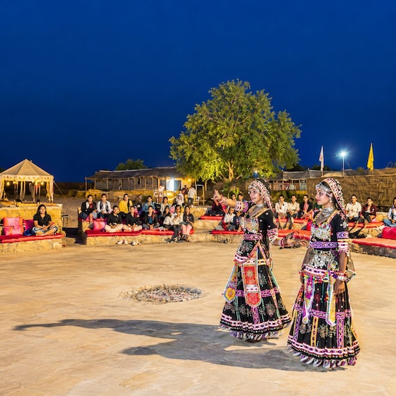 Two women in traditional attire performing a dance in front of an audience seated on red cushions. 2