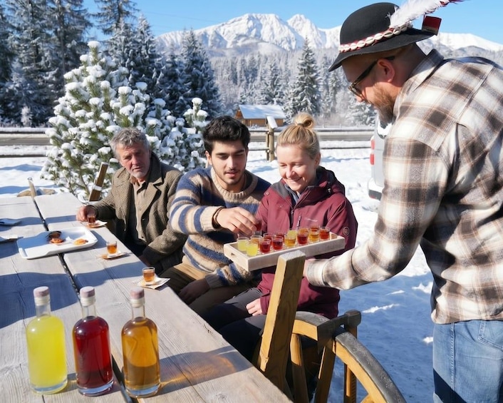 A group of people sitting at a wooden table outdoors with bottles of beverages and a tray of food. 2