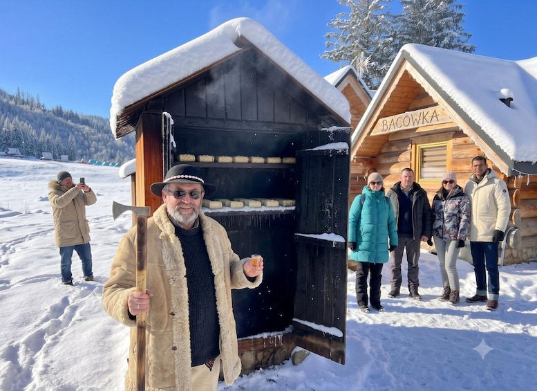 A wooden cabin with snow on the roof and a person holding a drink in front of it. 4