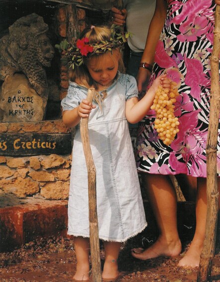 A child holding a bunch of grapes and a wooden stick, standing in front of a stone wall with a sign. 2