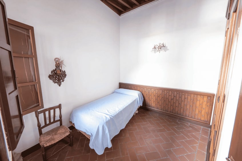 A small, clean room with a single bed, a wooden bench, a wall-mounted light fixture, and a door. 2