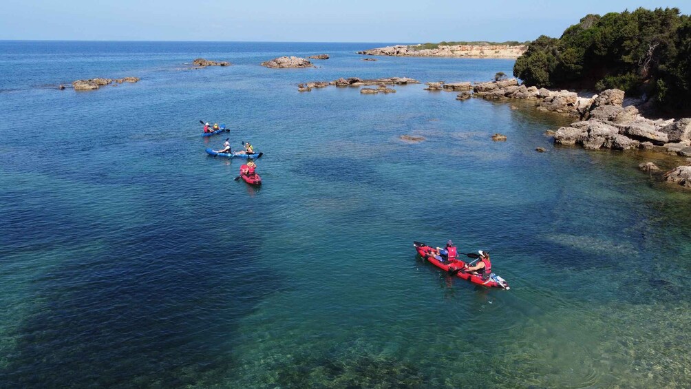 Kayakers in blue and red kayaks navigating a clear coastal waterway with rocky outcrops and a forested hill in the background. 1